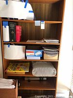 Front view of tall wooden bookcase with books, notebooks, and a box of envelopes on shelves.