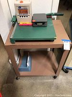 Brown metal office cart with a green paper cutter on top along with an electric scale and a small box.