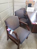 Photo showing two grey upholstered office conference chairs with wooden armrests placed near filing cabinets and an office desk.