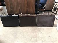 Photo showing three leather briefcases upright, displaying front and handles with visible wear and tear.