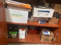 Photo of lower left cabinet shelf containing white plastic box, box of label sheets, HP ink cartridges, and small cardboard boxes
