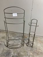 Two metal bathroom racks, one taller towel rack and one smaller toilet paper holder, placed on concrete floor against a wall.