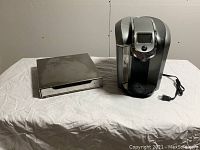 Keurig coffeemaker and metal pod holder side by side on a white cloth-covered surface.