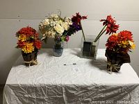 Image of four plastic flower arrangements on a white cloth, two wrapped in brown paper with raffia ties, one in clear glass vase, and one in blue and white ceramic vase.