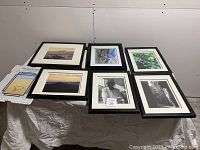 Six black wood picture frames laid out on a table, five contain photos or prints and one is an extra pane of glass.
