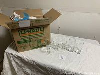 Wide view showing several tall and stemmed glasses arranged on white cloth nearby a U-Haul box for reference of lot packing.