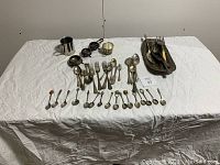 Full view of the silverware and collectible spoons assortment spread on a white cloth, including trays, tongs, spoons, forks, and small bowls.