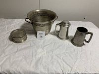 Wide photo showing all four pewter items on white tablecloth: champagne/wine bucket with handles, round coaster, and two beer steins, one smooth with lid and one hammered without lid.