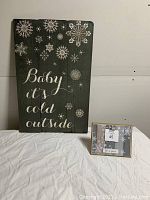 Photo of two wood winter decoration signs placed next to each other. Larger green wooden sign with snowflakes and text "Baby it's cold outside". Smaller framed sign with winter design and text.