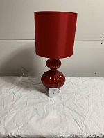 Full view of the red Bouclair table lamp with red lampshade on a white cloth surface against a white wall.
