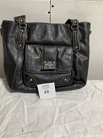 Front view of black Kenneth Cole Reaction leather tote bag, showing handles, front pocket with metal logo plate and textured leather surface.