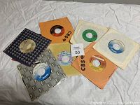 Photo of seven 45 RPM vinyl records with colored paper sleeves from labels CBS, Columbia, Capitol, and others, displayed on white fabric background.