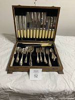 Full view of wood case open showing knives with ivory/cream handles lined on lid and forks, spoons, and serving utensils in bottom section on black velvet.
