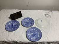 Wide view of three blue and white English transferware plates, clear glass teapot with lid, and antique cast iron bacon press on a white cloth background.