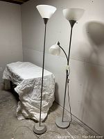 Pair of two freestanding floor lamps placed next to a covered table in a white-walled room.