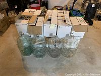 Four large clear glass wine carboys placed on the floor in front of stacked white cardboard cases containing wine bottles.