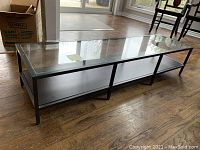 Rectangular coffee table shown from side angle on hardwood floor. Glass top and wood bottom shelf with black metal frame visible.