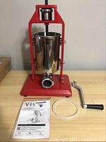 3L Sausage stuffer with red metal frame, stainless steel container, manual crank handle, rubber gasket, and instruction manual on light wood surface.