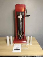 Front view of sausage stuffer with accessories including sausage tubes and instruction manual on a light wood table against grey wall