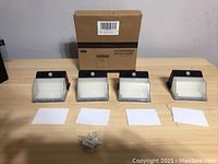 Set of four solar powered LED lights arranged in a row with mounting hardware visible in front, along with original packaging box in background.