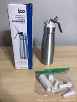 Photo of ICO whipped cream dispenser next to product box and plastic bag with accessories on wooden surface.