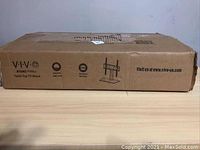 Closed cardboard box showing product details and brand Vivo with model STAND-TV003 printed on the box.