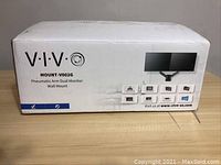 Photo of the VIVO MOUNT-V002G retail box showing the product name, images of the dual monitor mount, and website details.