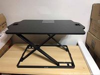 Front angle view of the black adjustable desk riser showing the X-frame lifting mechanism and surface handles.