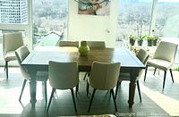 Eight beige upholstered dining chairs with ash wood legs arranged around wooden dining table near window