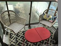 Two rattan chairs, one with a colorful cushion, two red stackable side tables on a black and ivory patterned rug on balcony floor.