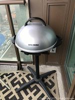 George Foreman grill with closed silver dome lid shown on black stand with four legs on balcony.