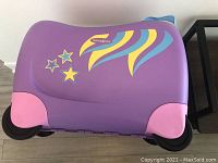 Top view of purple Samsonite ride-on suitcase showing colorful star and swirl designs, four wheels, and pink corner protectors.