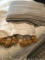 Cream and grey striped handwoven Moroccan blanket with large gold pom poms, folded underneath a grey textured knit blanket.
