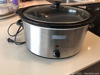 Side view showing oval stainless steel slow cooker with black plastic handles and a black dial control on front, glass lid on top.
