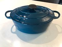 Full view of teal oval cast iron Dutch oven with lid and two loop handles on each side, placed on white surface.