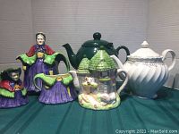 Four assorted teapots including vintage Old Lady with matching creamer and sugar bowl, green classic teapot, white floral teapot, and castle theme teapot shown together.