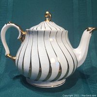 Front profile of the white ceramic teapot with gold swirl design and gold accents on the handle and lid finial.