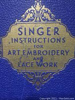 Close-up of the embossed gold title on the blue textured cover of the book, showing the words 'Singer Instructions for Art Embroidery and Lace Work'.