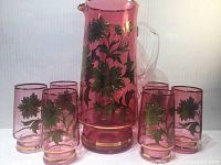 Full view of the purple glass pitcher with 6 matching tumblers arranged around it, showing gold floral pattern and clear handle on pitcher.