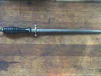 Full length view of the knife sharpening steel showing wooden handle, crossguard, and metal honing rod on wooden surface.