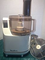 Front angled view of the white Moulinex food processor with clear plastic bowl and handle, accompanied by two stainless steel blades
