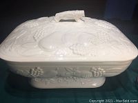 Exterior top and side view of creamy white ceramic vegetable warmer with detailed fruit and grapevine embossed design and handle on lid