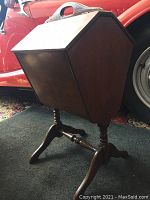 Front side view of wooden midcentury sewing/magazine stand showing squared compartment with hinged lid and handle at top mounted on spindle legs.