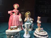 Photo showing all six pieces together on a green tablecloth including the pink lady figurine, two smaller figurines, the vase, and two trinket boxes.