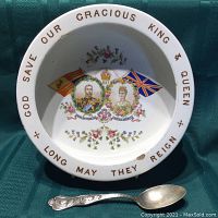 Top view of white ceramic bowl showing colorful portraits of King George V and Queen Mary with flags and floral decorations, and gold lettering around rim. Spoon lies inside bowl.