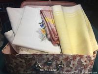 Seven assorted tablecloths partially folded inside floral case showing variety of colors and embroidery