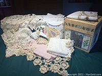 Photo showing a variety of lace table covers and doilies spread out with some folded linens on the side, displaying intricate crochet patterns in cream and white.