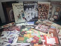 Photo showing an assortment of needlepoint and cross stitch pattern books including floral bookmark designs, nursery rhymes, and tissue box cover patterns.