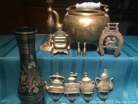 Full grouping of assorted brass items including teapot, shoehorn, vase, hooks, and smaller decorative brass pieces on blue textured fabric.