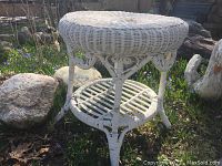 Full view of the white painted wicker round side table showing the two tiers and leg design in natural outdoor setting.